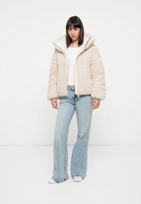 Beige fuzzy jas met een capuchon, voorzien van een voorkant rits en trekkoord, gedragen over een witte top en gecombineerd met lichtblauwe flared jeans.