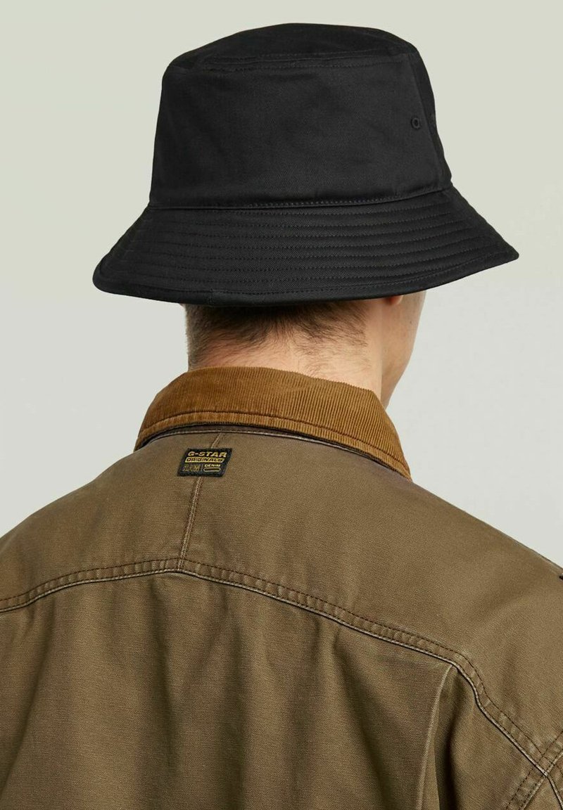 Chapeau seau en coton noir à large bord, associé à une chemise col en velours côtelé marron avec une étiquette au dos.