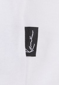 White fabric with a smooth texture features a black woven label displaying a white embroidered design.