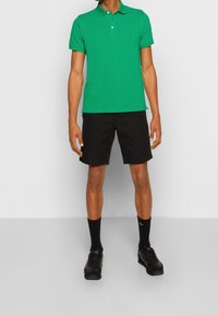 Zielona koszulka polo z kołnierzykiem, czarne szorty, czarne skarpetki i czarne sneakersy. Prosty design, bez wzorów, z minimalnym brandingiem na koszulce.