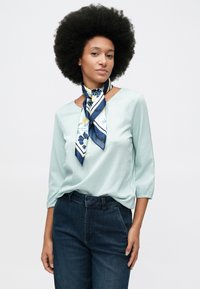 Γυναίκα με μεγάλο αφρο φορώντας μπλούζα με ανοιχτές ρίγες, σκούρα τζιν και μπλε μεταξωτό μαντήλι με λουλούδια δεμένο γύρω από τον λαιμό, που στέκεται μπροστά σε ανοιχτό φόντο.