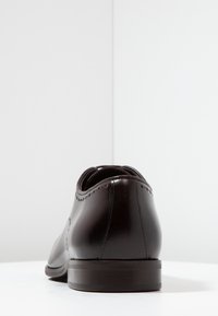 Chaussure habillée en cuir marron avec une finition lisse, bout arrondi, coutures décoratives le long des coutures, et une semelle basse et robuste de couleur marron.