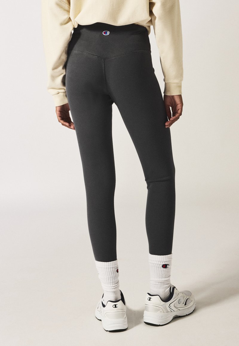 Leggings grigi in tessuto elasticizzato, aderenti, con una cintura alta decorata da un logo. Indossati con calzini bianchi e scarpe sportive.