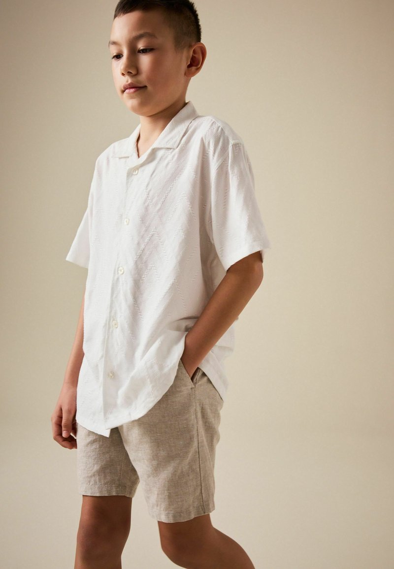 Niño de pie con las manos en los bolsillos, vistiendo una camisa blanca de manga corta abotonada y pantalones cortos beige claro contra un fondo liso.