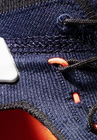 Detailní záběr na tmavě modrou tenisku s pletenou texturou. Obsahuje oranžové akcenty, černé tkaničky a bílé logo na boku.