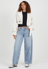 Przycięty beżowy trencz na czarnym topie, w połączeniu z szerokimi jasnoniebieskimi dżinsami i białymi trampkami w czarne paski.