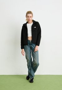 Black fleece zip-up jacket with hood, featuring a front logo patch and two side pockets, paired with blue jeans and black shoes.