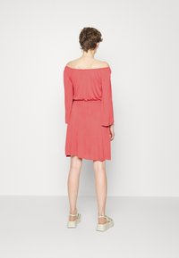 Vestido cor-de-rosa off-the-shoulder com mangas longas, cintura ajustada e saia rodada. Textura de tecido suave. Modelo usa sapatos de plataforma bege.