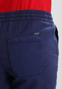 Námorně modré kalhoty s texturovanou tkaninou, elastickým pasem a jednou zadní kapsou. Má malou kovovou značku na kapse.