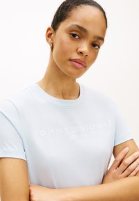 Ανοιχτό μπλε βαμβακερό μπλουζάκι με κοντά μανίκια και στρογγυλή λαιμόκοψη. Στο εμπρόσθιο μέρος εμφανίζεται το λογότυπο "TOMMY HILFIGER" με λευκό κείμενο.