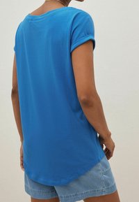 Blå kortærmet T-shirt med rund halsudskæring og sammenrullede ærmer, lavet af blødt stof, vist med denim shorts.