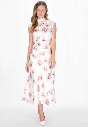 Kvinde iført en ærmeløs hvid midi-kjole med pink blomsterprint og høj halsudskæring, kombineret med hvide højhælede sko, mod en hvid baggrund.