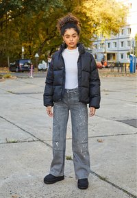 Chaqueta de plumas negra, blusa blanca con cuello alto, pantalones de mezclilla grises con salpicaduras de pintura y zapatos negros acolchados de tipo slip-on en un entorno urbano al aire libre.