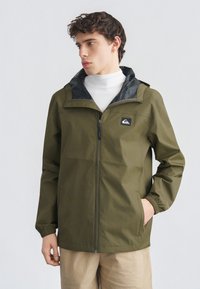 Νεαρός άνδρας που φοράει φούτερ με φερμουάρ σε ελιά πράσινο χρώμα και κουκούλα, λευκό ζιβάγκο και μπεζ παντελόνι, κοιτάζει αριστερά με το ένα χέρι στη τσέπη.