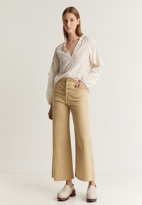 Blusa leggera di crema con maniche arricciate e piccoli pattern arancioni, abbinata a pantaloni beige chiaro a vita alta e gamba larga, e zoccoli di crema.