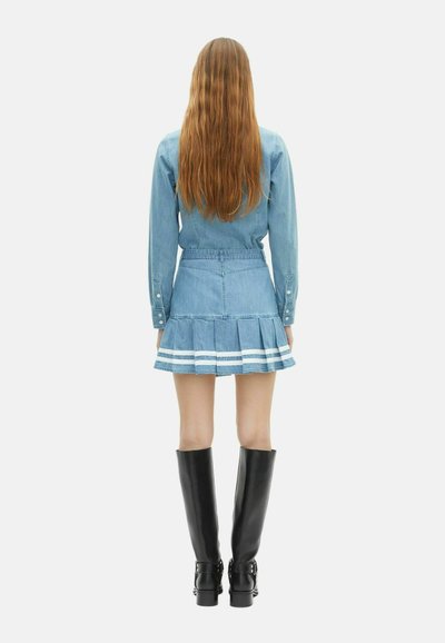 Vrouw met lang haar, gekleed in een blauwe denim jack en een geplisseerde rok met witte strepen, gecombineerd met zwarte kniehoge laarzen, staande met haar rug naar de kijker gekeerd.