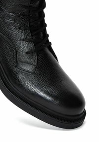 Botín negro con cordones, hecho de cuero texturizado, presenta una punta redonda y una suela de goma con un detalle de costura visible alrededor de la base.
