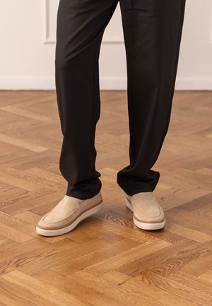 Benenden met zwarte broek en beige instapschoenen staand op een lichtbruine houten parketvloer.