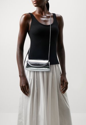 Femme portant un haut noir sans manches et une jupe plissée blanche, avec un petit sac bandoulière argenté à chaîne.