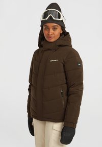Bruine pufferjack met horizontale stiksels, hoge kraag, twee zijzakken en logo. Draagt een donkere beanie en skibril.
