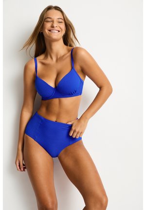 Kobieta uśmiechająca się i pozująca w teksturowanym niebieskim dwuczęściowym stroju kąpielowym na jednolitym jasnym tle.