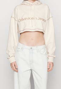 Felpa beige accorciata con cappuccio e cordoncini, dotata di logo in rilievo "Calvin Klein Jeans" e cuciture a contrasto. Abbinata a jeans azzurri chiari.