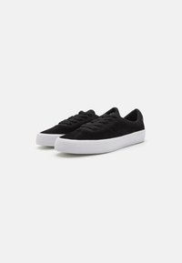 Adidași negri din suede cu talpă din cauciuc albă, vârf rotund și închidere cu șireturi. Partea superioară are o suprafață texturată și un design simplu.