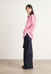 Long-sleeve, light pink striped shirt with button details, paired with dark, wide-leg jeans. Model stands against a neutral background.