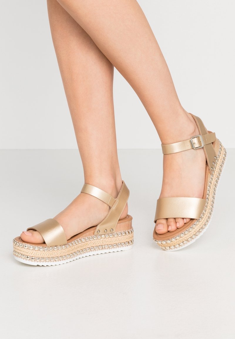 Füße, die goldene, metallische Plateausandalen mit Knöchelriemen tragen, mit Nieten um die geflochtenen Espadrille-Sohlen auf einem einfarbigen Hintergrund.