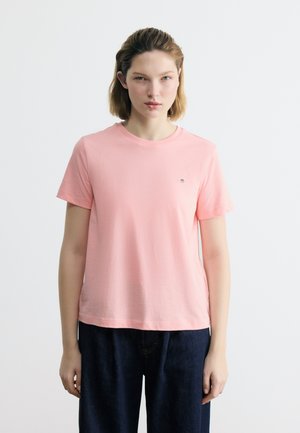 Νεαρή γυναίκα με κοντά μαλλιά, φοράει ένα απλό ροζ ανοιχτόχρωμο μπλουζάκι με κοντά μανίκια και σκουρόχρωμο παντελόνι, στέκεται μπροστά σε ένα λευκό φόντο.