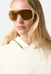 Białe, oversize okulary przeciwsłoneczne z brązowymi przyciemnianymi szkłami. Charakteryzują się kanciastym designem i wyrazistą oprawką. Dopełnione jasnym kapturem.