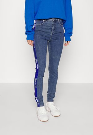 Personne portant un jean slim taille haute bleu avec des bandes latérales violettes et des baskets blanches, assorti à un haut bleu à manches longues.