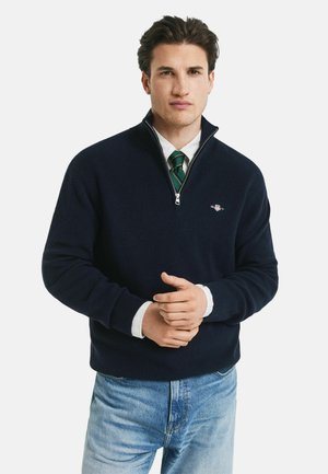 Man in een marineblauwe kwart-rits trui over een wit shirt en groene gestreepte stropdas, met lichtblauwe jeans, staande tegen een witte achtergrond.