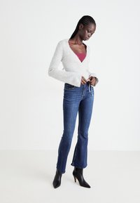 Hellgrauer Wickelpullover mit langen Ärmeln, kombiniert mit dunkelblauen Schlagjeans. Schwarze spitz zulaufende Absatzstiefel vollenden das Outfit.