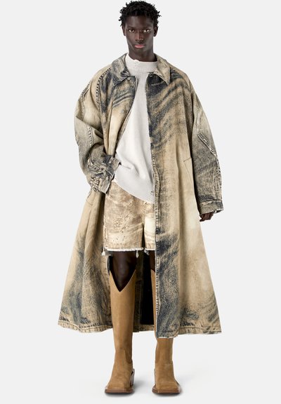 Personne grande portant un manteau ample beige et noir usé, un pull beige, un short assorti et des bottes hautes beige, debout devant un fond blanc.