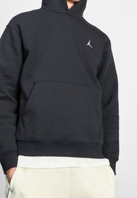 Schwarze Kapuzenjacke aus weichem Stoff, mit einer Kängurutasche und einem weißen Jordan-Logo auf der linken Brust. Glatte Textur, lockere Passform.