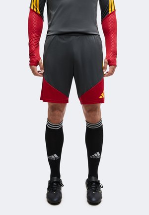 Athlet trägt dunkelgraue und rote Sportshorts, schwarze Adidas-Kniestrümpfe, schwarze Fußballschuhe und ein langärmeliges Oberteil mit roten Ärmeln und gelben Streifen.
