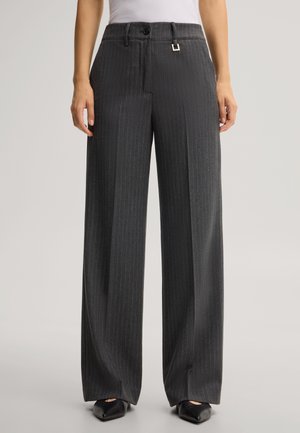 Frau trägt hoch taillierte, dunkelgraue Nadelstreifen-Weitbein-Hose mit Knopf- und Gürtelschlaufen-Detail, kombiniert mit schwarzen spitzen Schuhen.