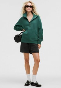Felpa verde con colletto alto e zip a metà, abbinata a pantaloni corti neri e mocassini. Gli accessori includono una piccola borsa rotonda nera.