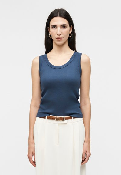 Femme aux longs cheveux foncés portant des boucles d'oreilles anneaux dorées, un haut sans manches bleu, un pantalon blanc et une ceinture marron, debout devant un fond uni.