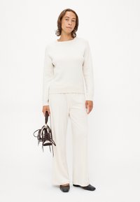 Sweater crema con cuello redondo, combinado con pantalones blancos holgados. El modelo sostiene un zapato marrón con cordones, situado sobre un fondo liso.