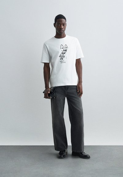 Muž oblečený v bílém tričku s grafikou Maison Kitsuné a volných tmavě šedých džínách, držící černou kabelku, stojící proti jednoduché světlé zdi.