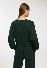 Ciemnozielona bluza z okrągłym dekoltem, z szerokimi rękawami i prążkowanym ściągaczem, w połączeniu z dopasowanymi spodniami z tego samego materiału i koloru.