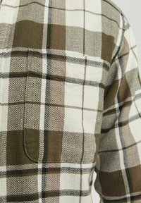 Cămașă în carouri, cu nuanțe de maro, crem și negru, realizată dintr-un material texturat, având un singur buzunar la piept. Designul include pătrate de dimensiuni variate.