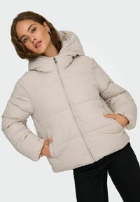 Giacca a vento avvolgente beige con cappuccio, cuciture orizzontali, chiusura frontale con zip e tasche laterali, indossata su pantaloni neri. Texture morbida e imbottita.