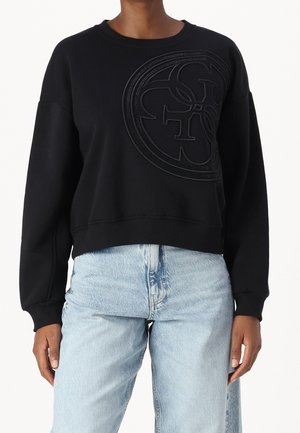 Persoon die een zwarte sweatshirt draagt met een groot reliëf rond logo op de borst, gecombineerd met lichtblauwe spijkerbroek.