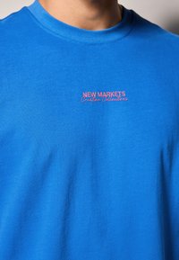 Blå bomulds t-shirt med rund hals. Centralt placeret pink broderi med teksten "NEW MARKETS Creative Collectives." Glat tekstur.