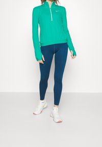 Žena oblečená v tyrkysové sportovní mikině na zip, námořnických legínách s kapsami a bílých teniskách se žlutým logem Nike, stojící na šedé podlaze.