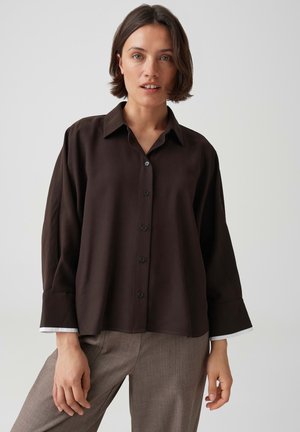 Donna con capelli corti castani che indossa una camicia larga marrone scuro con bottoni e pantaloni marroni chiari su sfondo uniforme.