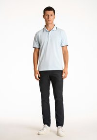 Camisa polo de color azul claro con cuello, ribete negro y logo en el pecho; combinada con pantalones oscuros y zapatillas blancas.